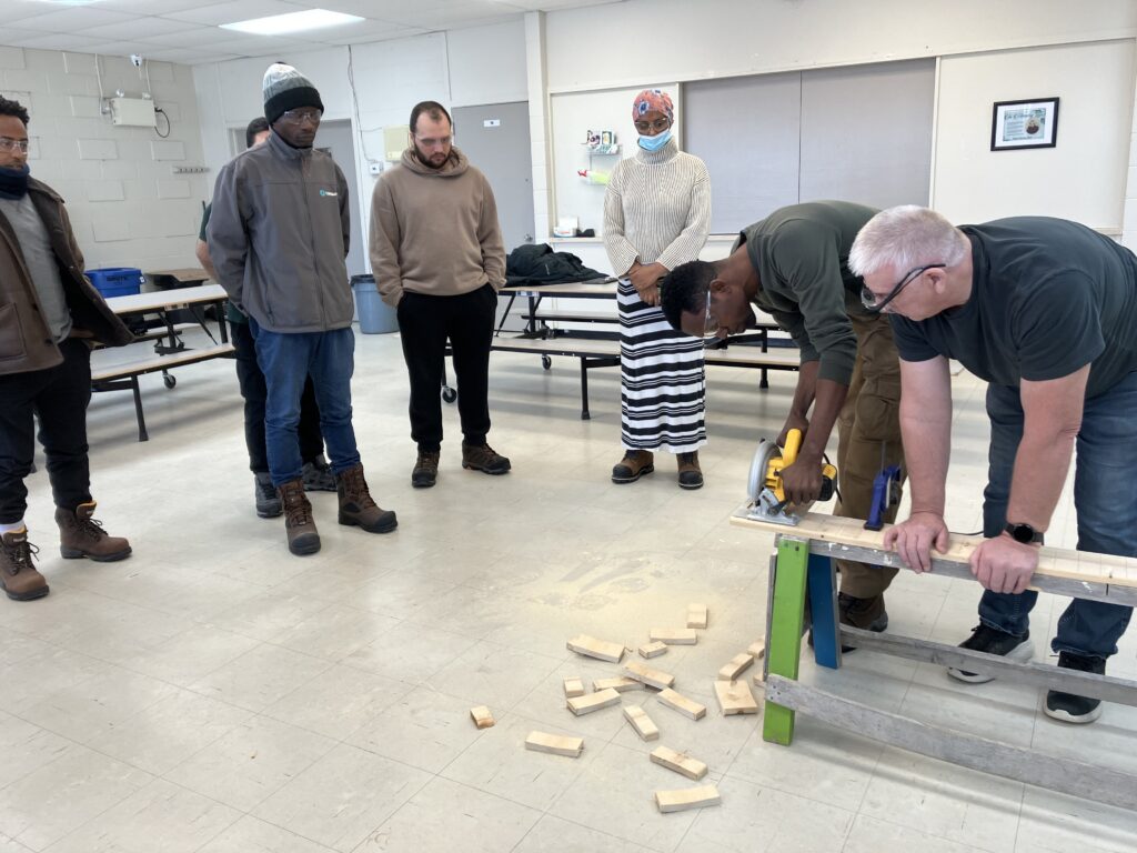 Participants in the Build Your Future construction training program practice using a circular saw under instructor supervision while other newcomers observe the hands-on woodworking demonstration.