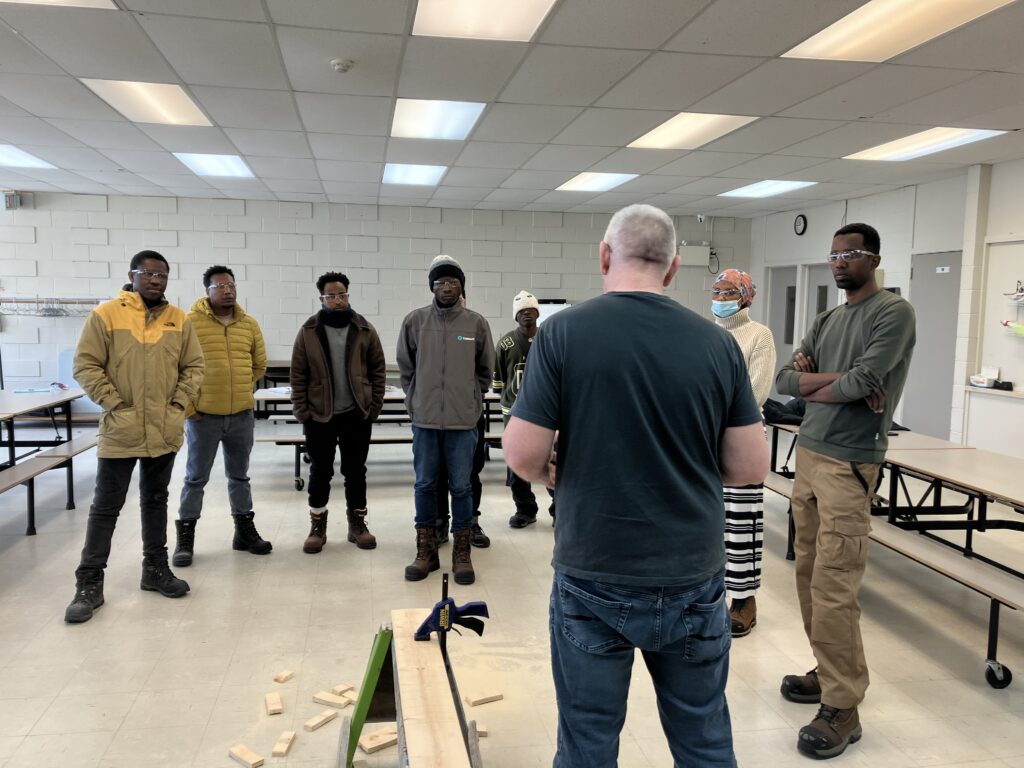Instructor demonstrates construction techniques to a diverse group of newcomers enrolled in the Build Your Future program, showing hands-on skills training in a workshop setting.