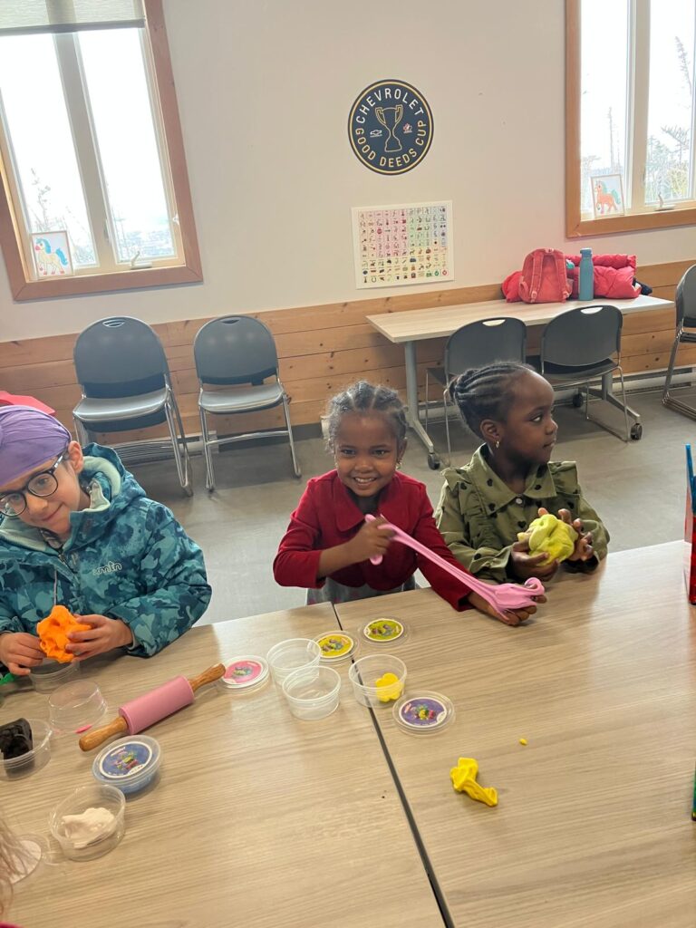 Three HIPPY program children doing play-dough crafts at table with Chevrolet Good Deeds Cup sign in background