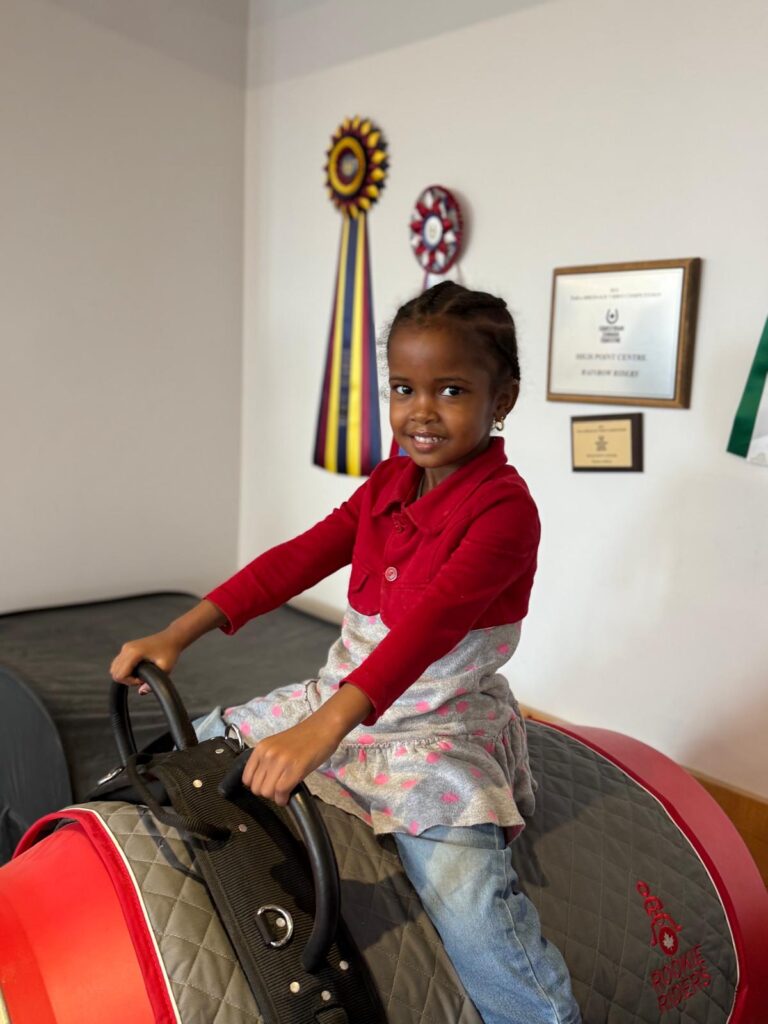 Young girl in red cardigan smiling while sitting on saddle at Rainbow Riders NL