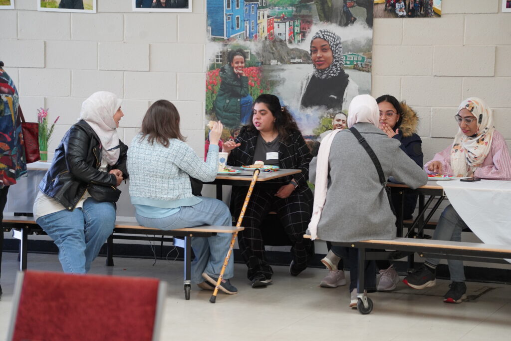 An audience of diverse adults and youth sits attentively in rows of red chairs and benches inside a brightly lit room. 