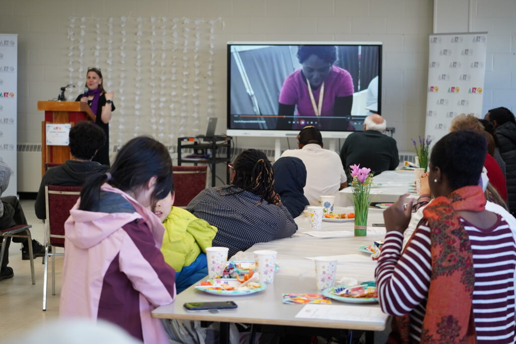 A diverse group of volunteers gathers around long tables covered with white tablecloths, engaging in conversation and reviewing documents. Some wear name tags; others hold papers or cups. Pink artificial flowers in green vases decorate the tables. 