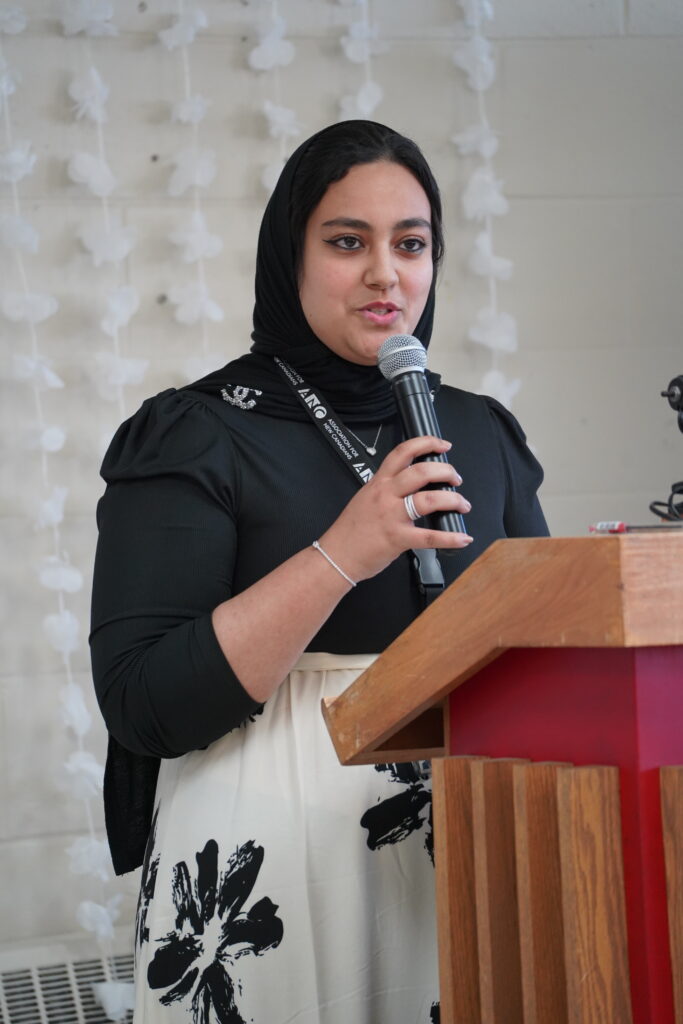A woman wearing a black hijab and a black-and-white floral dress stands at a wooden podium, speaking into a handheld microphone. She wears a lanyard with “ANC – Association for New Canadians” printed on it. Behind her, white paper flowers hang in vertical strands against a light-colored wall. Her expression is engaged and confident as she addresses the audience.